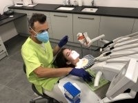 Widok na gabinet stomatologiczny podczas pracy lekarza. Wnętrze jest wyposażone w ciemne meble medyczne z czarnym blatem, które kontrastują z jasnym fotelem dentystycznym i białymi ścianami.