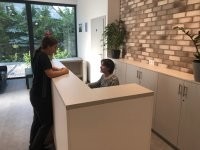 Strefa recepcji z minimalistyczną białą ladą. Recepcjonistka rozmawia z pacjentką. Charakterystycznym elementem wnętrza jest ozdobna ściana z jasnej cegły oraz duże przeszklenia wpuszczające naturalne światło.