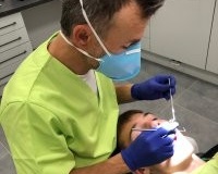 Stomatolog w zielonym stroju medycznym, maseczce i rękawiczkach wykonuje zabieg u pacjentki. Widoczna profesjonalna lampa zabiegowa oraz nowoczesna, biała aparatura dentystyczna.