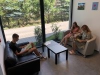 Jasna i nowoczesna poczekalnia. Trzech pacjentów siedzi na wygodnych fotelach i sofie przy czarnym stoliku kawowym, przeglądając czasopisma. Przez duże, przeszklone ściany widać zieleń na zewnątrz.