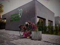 Nowoczesny, parterowy budynek gabinetu o ciemnej elewacji. Przed wejściem stoi rower z koszami pełnymi kolorowych kwiatów, a obok widoczna jest duża donica z roślinami na tle przeszklonych drzwi.